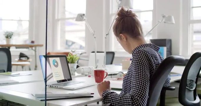 Young business woman working at shared desk in trendy hipster start up office using big data on laptop computer