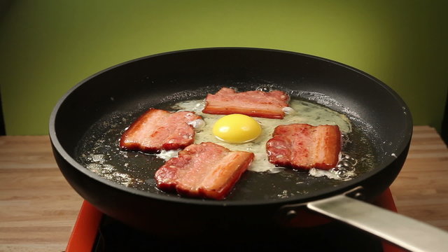 Close-up of cooking fried eggs for breakfast. An egg being dropped in a frying pan with bacon.