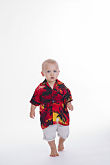 A young blonde male toddler in a colorful shirt and shorts is walking towards the camera.
