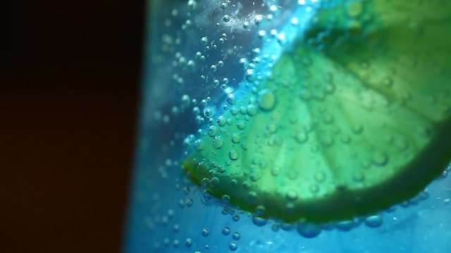 Color footage of a slice of lemon in a glass of soda, with ice cubes and some blue syrup.