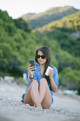 Young woman sitting at the beach drinking coffee and using her mobile phone
