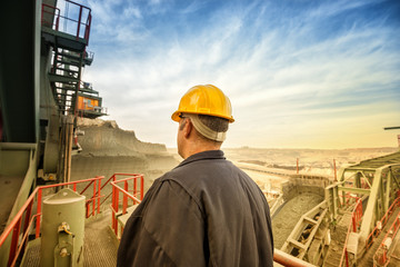 Coal mine engineer with a helmet standing on a huge drill machine and looking at digging site. Rear view. Beautiful and colorful sky in background.