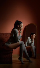 Attractive sexy brunette in white comfortable pullover posing sitting on a sofa indoors. Portrait of sensual young woman posing in front of large mirror. Beautiful girl with long stockings in her room