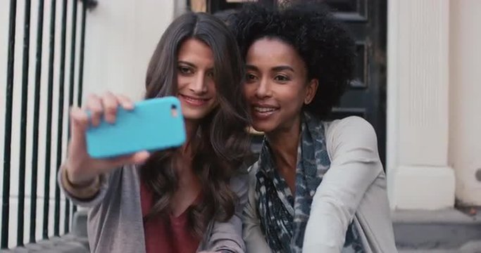 Two beautiful woman friends sitting on steps having fun talking selfie on smart phone