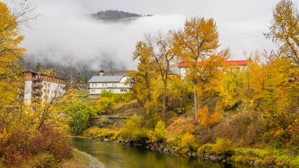 Fototapeta na wymiar River bank in the mist, autumn forest on the riverside, beautiful trees and small houses on a background of mountains