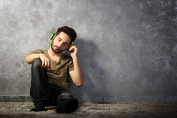 Young bearded man listens music with headphones sitting on grey wall background