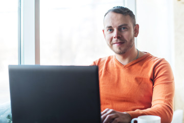 Fototapeta premium Handsome young man working on notebook, smiling, looking at camera, while enjoying coffee in cafe