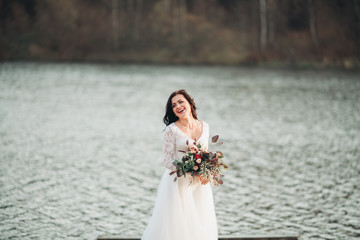 Fototapeta na wymiar Gorgeous brunette bride in elegant dress holding bouquet posing near forest and lake