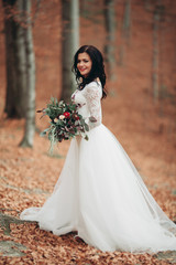 Gorgeous brunette bride in elegant dress holding bouquet  posing near forest and lake