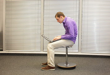 text neck - man in slouching position sitting on pneumatic stool, working with laptop in his office