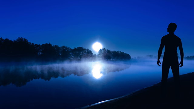 The man stand on the river bank on a background of light of the moon. Time lapse. Wide angle