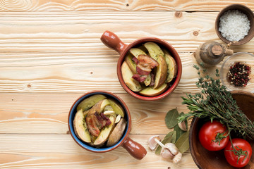 fried potatoes and bacon in ceramic forms on wooden table with clay plate with tomatoes,  thyme, rosemary, sea salt, olive oil in clay jug, garlic,  pepper, bay leaf , top view