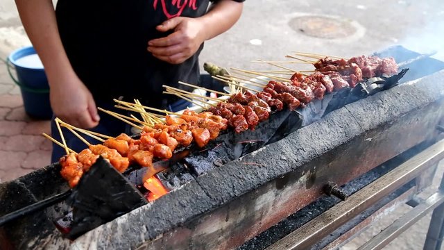 Vendor preparing delicious barbecue chicken and beef satay on charcoal grille with shallow focus on center skewers 