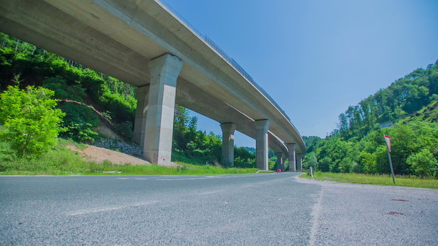Small vintage car is driving on a country road that goes under a highway. 