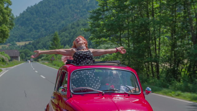 An older lady stands up and puts her hands up during a car ride in a red yugo
