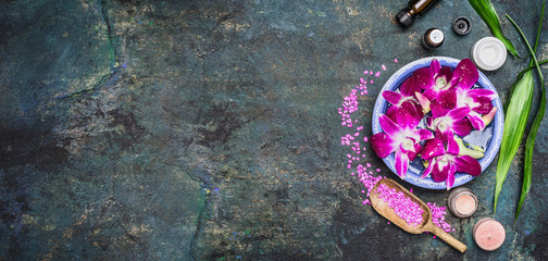 Spa setting with water bowls , pink orchid flowers ,sea salt, cosmetic cream and essential oil on dark rustic background , top view, place for text, horizontal, banner. Wellness concept