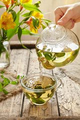 Peppermint tea pouring in glass cup from teapot, yellow flowers on vintage wooden background....