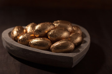 Close-up of Easter eggs in gold foil in a heart shaped wooden bowl against a dark background. 