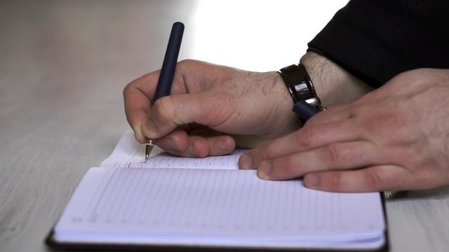 Close shot of a human hand writing something on the note paper on the foreground.