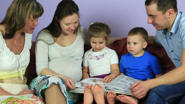 Parents with kids and grandmother consider an album with photos. Happy family waiting for third child.