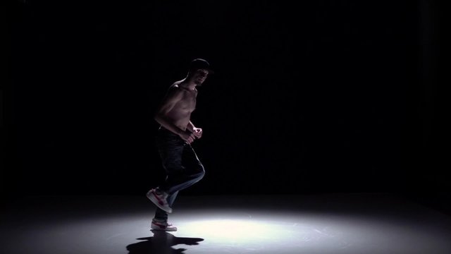 Breakdance dancer man in trousers with naked torso goes on dance on black, shadow, slow motion