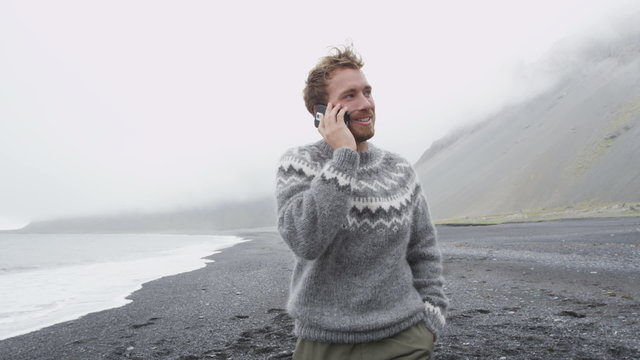 Smart phone man talking on smartphone walking on black sand beach on Iceland wearing Icelandic sweater by the ocean sea. Handsome Caucasian male model using smartphone. 60 FPS.