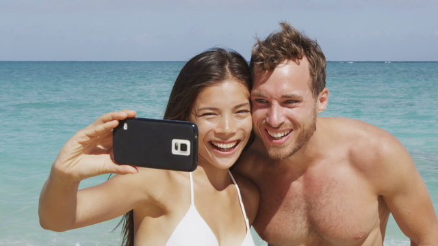 Phone selfie beach holiday couple having fun in love laughing. People taking photo with smartphone using smart phone camera. Young beautiful multicultural Asian Caucasian couple taking self portrait.