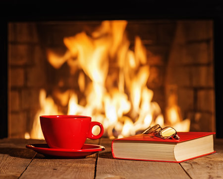 Red cup, glasses and old book near fireplace on wooden table.