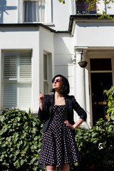 Girl in black dress with polka dots and sunglasses standing on the streets of London. Holding a cigarette.