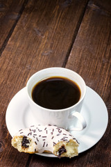 Coffee cup is placed on a wooden board. The half of filled american donut is lying on the saucer. Photo is edited as an vintage with dark edges.