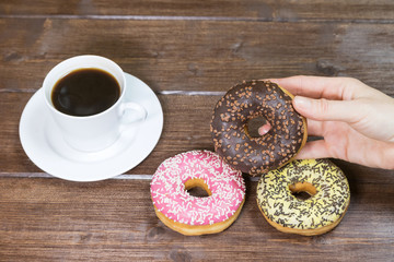 Two American donuts and cup of coffee are lying on a wooden table. Female hand is holding the third chocolate donut.