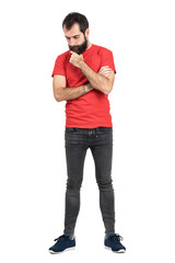 Young hipster in red t-shirt stroking beard white thinking and looking down. Full body length...