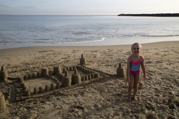 Cute blond girl standing by her sand castle smiling