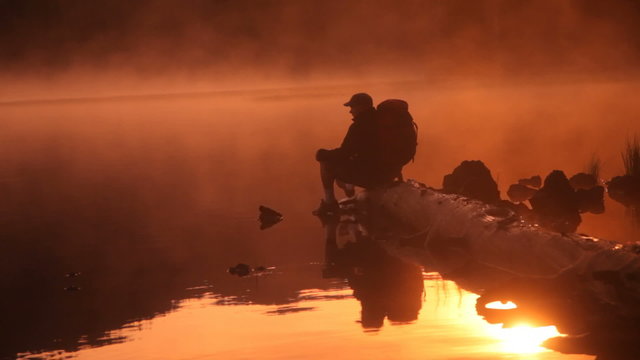 Backpacker at sunrise sitting on log in lake