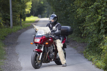 Naklejka premium Motorcyclist lost on the road through the forest looking at map