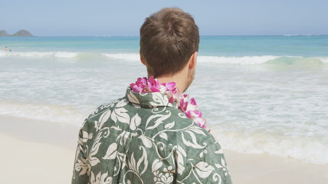 Young man standing on Hawaiian beach. Rear view of male tourist wearing flower lei garland and Aloha clothing. Visitor is looking at ocean during vacation on sunny day.