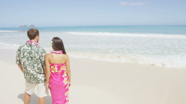 Young couple standing on Hawaiian beach. Rear view of tourists wearing flower lei garlands and Aloha clothing. Visitors are looking at beautiful sea during vacation on sunny day.