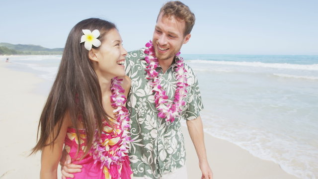 Happy couple walking with arms around on Hawaiian beach. Visitors are wearing flower lei garlands and Aloha clothing. Young tourists are spending leisure time on sunny day.