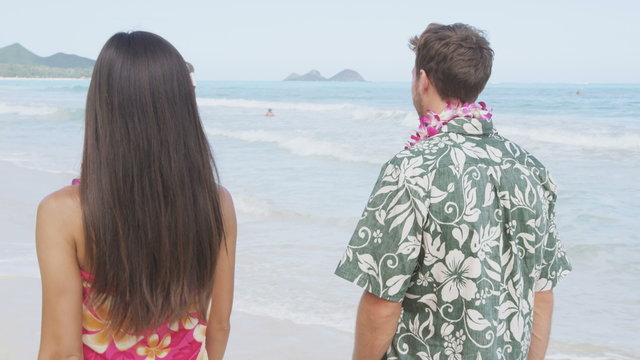 Happy couple in Aloha clothing walking on Hawaiian beach. Rear view of man and woman spending leisure time on sea shore. Visitors are enjoying their vacation on sunny day.