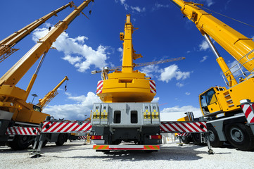 Mobile construction cranes with yellow telescopic arms and big tower cranes in sunny day with white clouds and deep blue sky on background, heavy industry  - 105220052