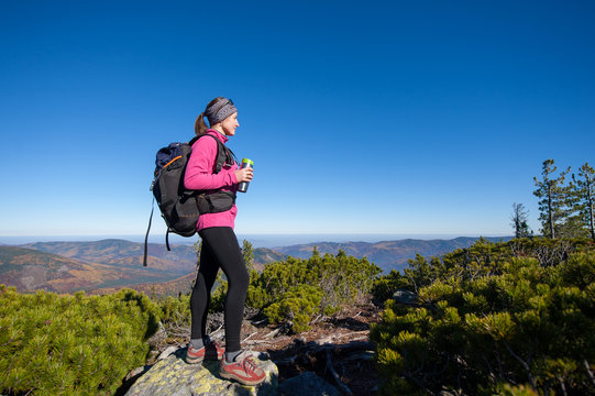 Female backpacker reaching her goal standing on peak of the mountain, holding thermos and enjoying the beautiful landscape. Sunny autumn time.