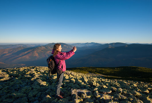 Young beautiful girl standing on rocky mountain top and taking picture with her smartphone. Beautiful mountains on the background. Ecotourism and healthy lifestyle concept. Sunny fall day.