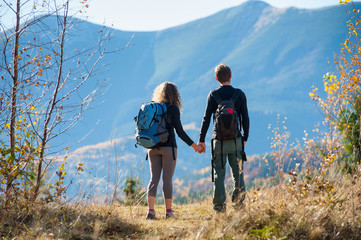 Fototapeta na wymiar Rear view of young backpackers couple looking into distance enjoying the overlook view and holding hands on a warm sunny autumn day in the mountains. Mountain on the background. Warm fall time.