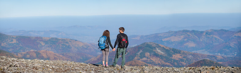 Rear view of young couple hikers with backpacks standing on the ridge of the mountain, enjoying the panorama view of beautiful open overlook on the mountains. Couple is holding hands. Wide angle view