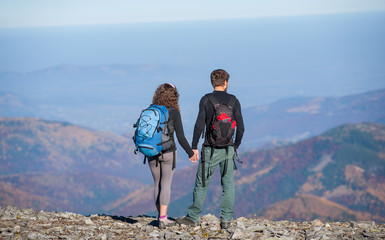 Rear view of young couple tourists with backpacks standing on the ridge of the mountain, enjoying the view of beautiful open overlook on the mountains. Couple is holding hands. Sunny autumn day.