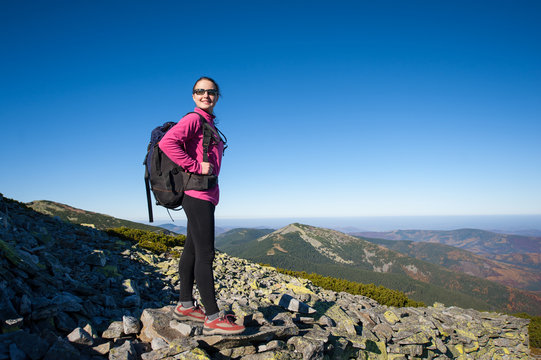 Young female backpacker standing on rocky mountain peak smiling and looking to the camera. Fall.
