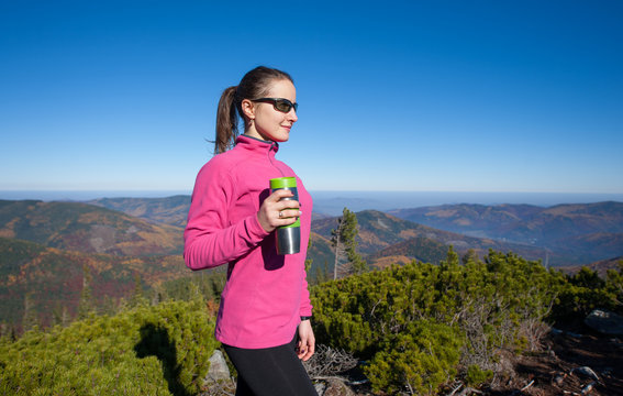 Young athlete woman hiker reached the beautiful mountain peak, enjoying her rest and drinking tea/coffee out of thermos. Woman is wearing pink jacket and sunglasses. Warm autumn time.