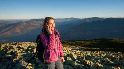 Fototapeta premium Portrait of young cute female backpacker standing on rocky top of the mountain, smiling, looking to the sun and talking cell phone. Side view. Healthy lifestyle concept. Sunny autumn evening.