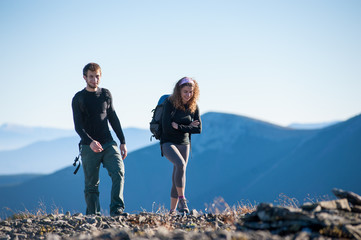 Portrait of male and female hikers walking up in the mountains. Front view with beautiful mountains on the background. Girl is smiling and looking down, guy is looking into the camera.