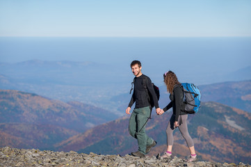 Active couple of tourists climbing up with backpacks on the ridge of the mountain, open overview on the mountains on the background. Couple is smiling and holding hands. Sunny autumn day. Side view.